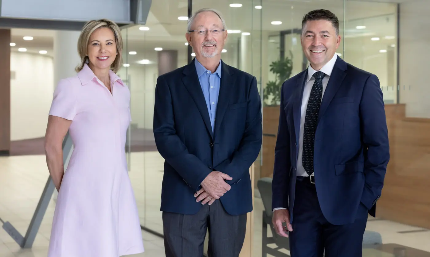 Three professionals posing together in a modern office setting, showcasing a confident and approachable atmosphere. The woman on the left wears a light pink dress, while the two men are attired in business suits, one in navy blue and the other in a dark suit with a patterned tie. The background features glass walls and a well-lit interior, emphasizing a professional environment.