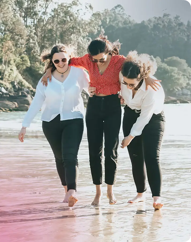 Three women walking barefoot on a beach, enjoying a sunny day. They are dressed in casual outfits, with one wearing a red polka dot top and sunglasses. The background features lush greenery and a serene ocean view, capturing a moment of friendship and relaxation.