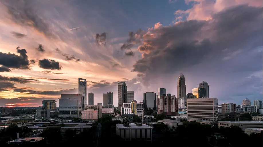 Panoramic view of the Charlotte skyline during sunset, showcasing modern skyscrapers against a dramatic sky filled with clouds. The image captures the vibrant urban landscape and the warm colors of the setting sun, highlighting Charlotte's architectural diversity.